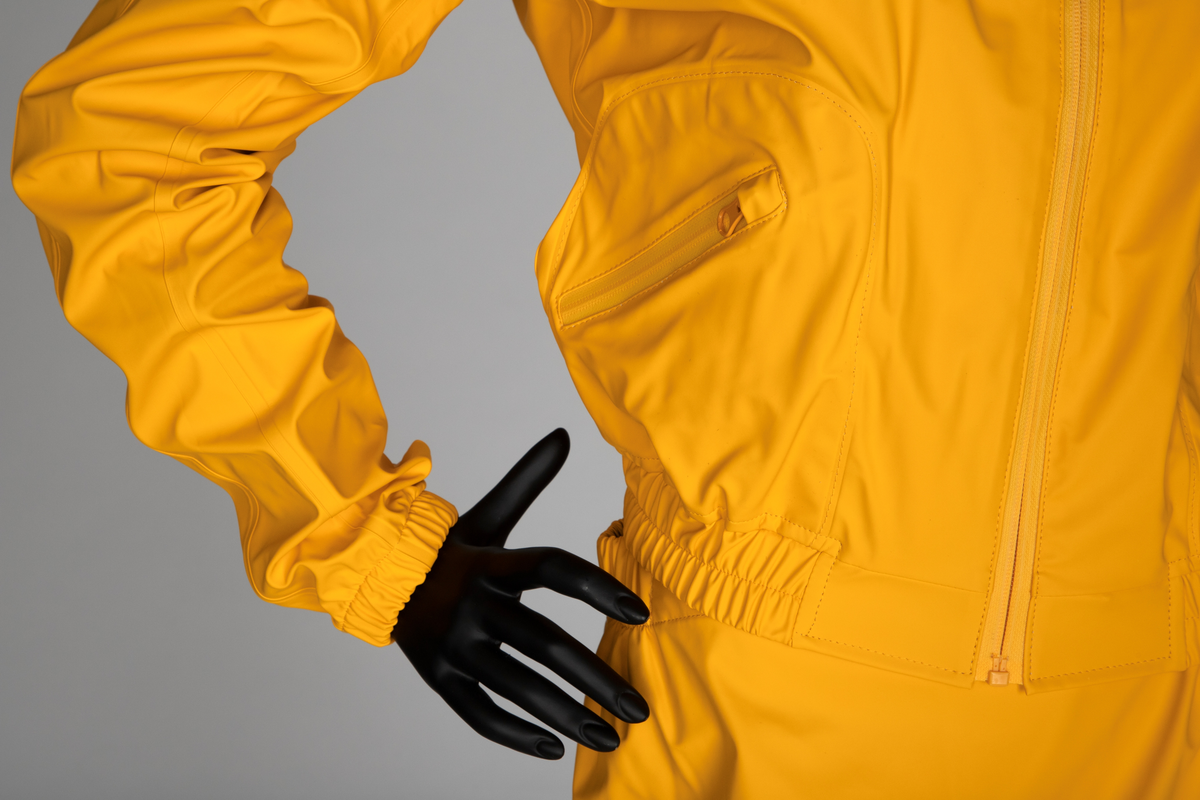 Drakt som består av battlejakke og omslagsskjørt i gult regntøy. Jakken har frontlukning med enveisglidelås og linning i halsen lukket med trykknapp. Skråstilt lomme, lukket med glidelås nederst på hvert forstykke. Jakken har innsydd strikk i nedre kant og isydde, tosøms-ermer og bærestykke. Ermer og bole er avsluttet med isydd elastikk i nederkant. Dette gir en romslig blusong-effekt. Alle viktige sømmer er både sydd og sveiset. Knehøyt omslagsskjørt med åpning midt bak, isydd strikk i livet og knepping med to trykknapper der skjørtet har sin circa 10cm's overlapping bak. Skjørtet er klippet i ett stykke og kantet i nederkant med påsydd skråbånd.