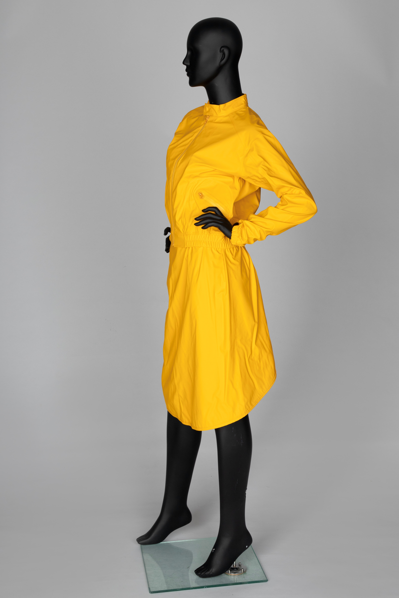 Drakt som består av battlejakke og omslagsskjørt i gult regntøy. Jakken har frontlukning med enveisglidelås og linning i halsen lukket med trykknapp. Skråstilt lomme, lukket med glidelås nederst på hvert forstykke. Jakken har innsydd strikk i nedre kant og isydde, tosøms-ermer og bærestykke. Ermer og bole er avsluttet med isydd elastikk i nederkant. Dette gir en romslig blusong-effekt. Alle viktige sømmer er både sydd og sveiset. Knehøyt omslagsskjørt med åpning midt bak, isydd strikk i livet og knepping med to trykknapper der skjørtet har sin circa 10cm's overlapping bak. Skjørtet er klippet i ett stykke og kantet i nederkant med påsydd skråbånd.