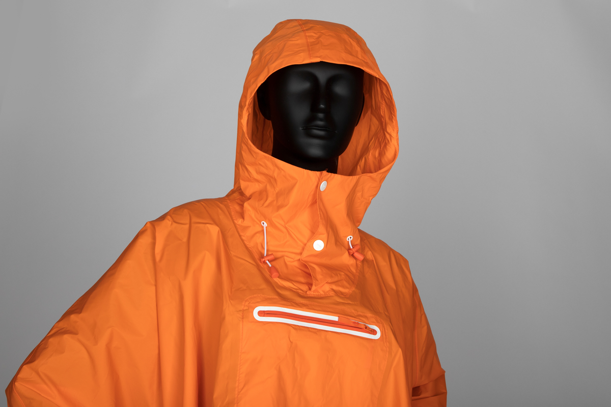Oransje regnponcho med hette. Ponchoen har en løsthengende sirkelform. Trykknapper nederst til ermene og hvit skråkant på nederkanten. Hette med åpning og to hvite trykknapper. Snor og maljer med snorstopper og snorregulerer. Midt foran på brystet sveiset glidelås, markert med hvit kant.