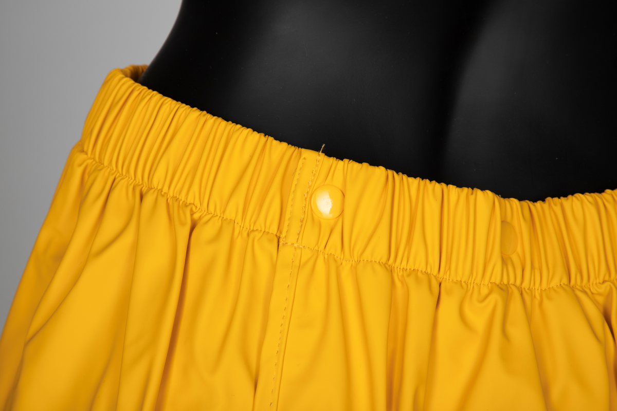 Drakt som består av battlejakke og omslagsskjørt i gult regntøy. Jakken har frontlukning med enveisglidelås og linning i halsen lukket med trykknapp. Skråstilt lomme, lukket med glidelås nederst på hvert forstykke. Jakken har innsydd strikk i nedre kant og isydde, tosøms-ermer og bærestykke. Ermer og bole er avsluttet med isydd elastikk i nederkant. Dette gir en romslig blusong-effekt. Alle viktige sømmer er både sydd og sveiset. Knehøyt omslagsskjørt med åpning midt bak, isydd strikk i livet og knepping med to trykknapper der skjørtet har sin circa 10cm's overlapping bak. Skjørtet er klippet i ett stykke og kantet i nederkant med påsydd skråbånd.