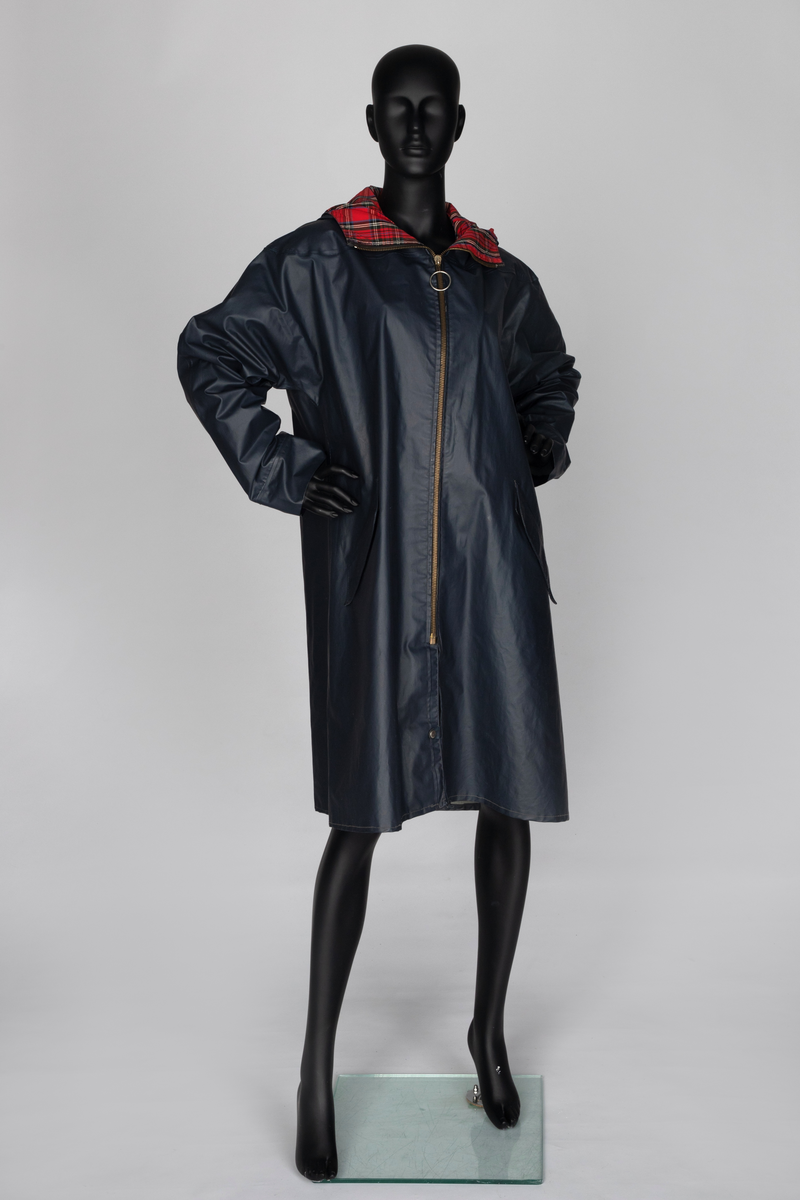 Lang regnkåpe med hette. Marineblå med rødrutet fôr i krage og hette og blått belegg ned langs åpningen. Matt PVC-overflate. Har nok hatt trekksnor i hetten, men den er ikke der nå. Tosøms-ermer, skåret iett med for- og bakstykke og sydd og sveiset. Helskåret bakside. Skråstilte stikklommer i hoftehøyde på hvert forstykke, med klaff, og forsterkninger i åpningen. Opphengshempe i nakken. Lukket midt foran med kraftig metallglidelås med stor ring. Glidelåsen stopper 28cm ovenfor nederkanten, men åpningen nedenfor lukkes midtveis med trykknapp. Trekantet, åpent bærestykke bak, til ventilering. Nedre snipp er kneppet fast, men kan kneppes opp og avdekker da refleks på innsiden av klaffen.