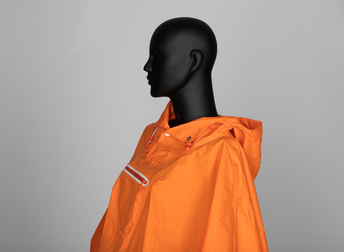 Oransje regnponcho med hette. Ponchoen har en løsthengende sirkelform. Trykknapper nederst til ermene og hvit skråkant på nederkanten. Hette med åpning og to hvite trykknapper. Snor og maljer med snorstopper og snorregulerer. Midt foran på brystet sveiset glidelås, markert med hvit kant.