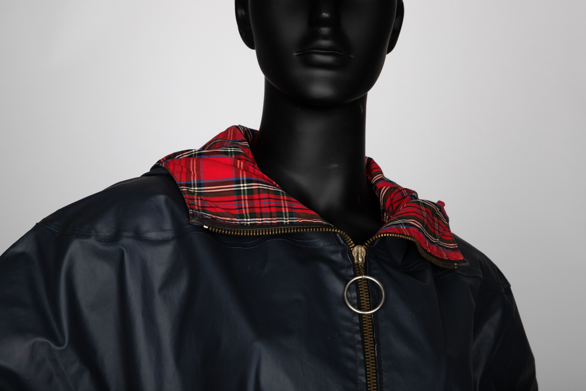 Lang regnkåpe med hette. Marineblå med rødrutet fôr i krage og hette og blått belegg ned langs åpningen. Matt PVC-overflate. Har nok hatt trekksnor i hetten, men den er ikke der nå. Tosøms-ermer, skåret iett med for- og bakstykke og sydd og sveiset. Helskåret bakside. Skråstilte stikklommer i hoftehøyde på hvert forstykke, med klaff, og forsterkninger i åpningen. Opphengshempe i nakken. Lukket midt foran med kraftig metallglidelås med stor ring. Glidelåsen stopper 28cm ovenfor nederkanten, men åpningen nedenfor lukkes midtveis med trykknapp. Trekantet, åpent bærestykke bak, til ventilering. Nedre snipp er kneppet fast, men kan kneppes opp og avdekker da refleks på innsiden av klaffen.