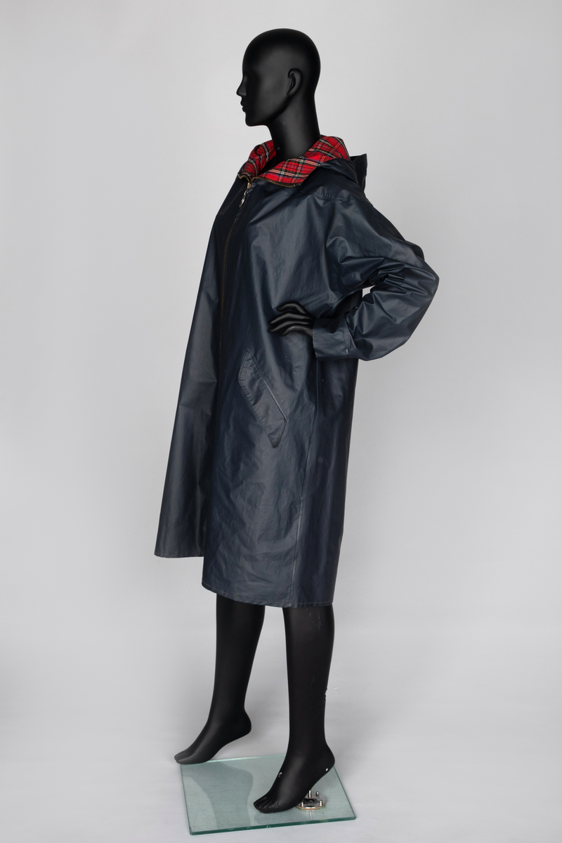 Lang regnkåpe med hette. Marineblå med rødrutet fôr i krage og hette og blått belegg ned langs åpningen. Matt PVC-overflate. Har nok hatt trekksnor i hetten, men den er ikke der nå. Tosøms-ermer, skåret iett med for- og bakstykke og sydd og sveiset. Helskåret bakside. Skråstilte stikklommer i hoftehøyde på hvert forstykke, med klaff, og forsterkninger i åpningen. Opphengshempe i nakken. Lukket midt foran med kraftig metallglidelås med stor ring. Glidelåsen stopper 28cm ovenfor nederkanten, men åpningen nedenfor lukkes midtveis med trykknapp. Trekantet, åpent bærestykke bak, til ventilering. Nedre snipp er kneppet fast, men kan kneppes opp og avdekker da refleks på innsiden av klaffen.