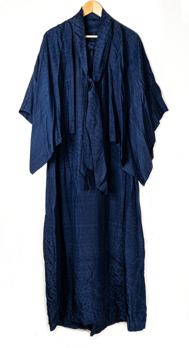 Klänning med tillhörande cape. Invävt mönster av små blommor. Klänningen pryds med en rosett vid halslinningen framtill. Lätt utställda ärmar. Capen knyts fram med ett brett band.