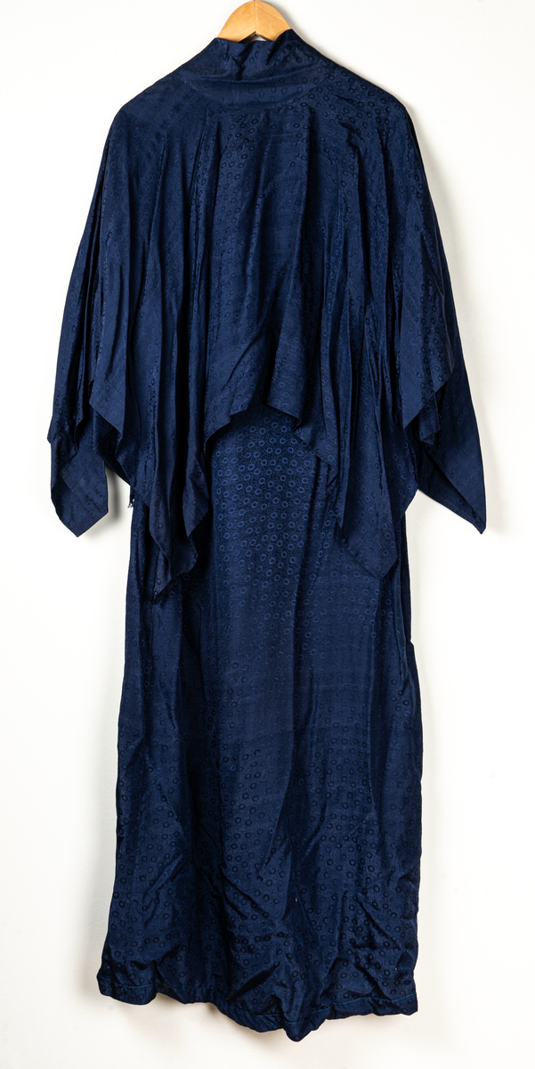 Klänning med tillhörande cape. Invävt mönster av små blommor. Klänningen pryds med en rosett vid halslinningen framtill. Lätt utställda ärmar. Capen knyts fram med ett brett band.