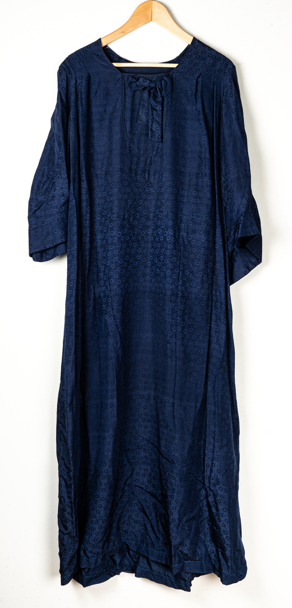 Klänning med tillhörande cape. Invävt mönster av små blommor. Klänningen pryds med en rosett vid halslinningen framtill. Lätt utställda ärmar. Capen knyts fram med ett brett band.