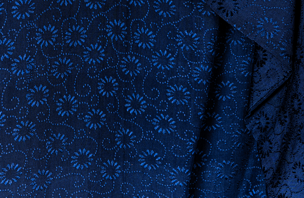 Klänning med tillhörande cape. Invävt mönster av små blommor. Klänningen pryds med en rosett vid halslinningen framtill. Lätt utställda ärmar. Capen knyts fram med ett brett band.