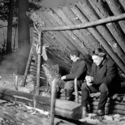 Gapaskjul i Norsk Skogmuseums friluftsmuseum på Prestøya i Elverum i Hedmark, fotografert i 1962, antakelig under opptakene til dokumentarfilmen «Liv og husvær i skogen». Et gapaskjul var et krypinn som skulle gi et visst vern mot klimaet. Det besto i sin enkleste form av et skråtak – pulttak – med enkle, trekantete sidevegger som også gav et visst vern mot vinden. Slike skjul ble vanligvis lagd av skrapvirke fra skogen der de ble bygd. Dette eksemplaret består av en brisk, lagd av paralleltliggende smågran (staurer med 5-8 centimeters diameter) i drøyt 2 meters bredde og 3 meters lengde. Når slike gapaskjul ble brukt i skogen og langs fløtingsvassdragene ble det naturligvis lagt granbar på briskene, for å skape et noe mjukere sitte- og liggeunderlag enn staurene. Ved hjørnene i den fremre enden av brisken er det reist  to stolper med kløftet topp, som er drevet ned i bakken. Mellom de nevnte kløftene er det lagt en horisontal stokk, en ås. Fra denne stokken og mot den bakre enden av brisken er det lagt et pulttak av halvkløvninger. Sideveggene i gapaskjulet er lagd etter et skigardsprinsipp, med staurer som er drevet ned i bakken parvis og mellomliggende staur, antakelig holdt på plass ved hjelp av gransveig. All stauren i sideveggene har rotenden fram, slik at vegghøyden avtar bakover, slik takhellinga også gjør. da dette fotografiet ble tatt satt to arbeidkledde menn på ytterkanten av brisken i gapaskjulet. Ved sida av den ene sto det en neverkont. Karene satt i lyset fra en nying. Dette var stokker som var lagt parallelt, over hverandre med et pyramidalt tverrsnitt, her med to stokker i den nestee floa og en oppå. For å holde det hele på plass ble det stukket kjepper – «haldmerrer» – ned i bakken på begge sider ved nyingens ytterkanter. Yteveden på stokkene ble gjerne litt oppfliset ved hjelp av ei øks, noe som gjorde dem lettere antennbare. Nyingen kunne gi langvarig lys og varme til dem som kvilte og sov i gapaskjulet.