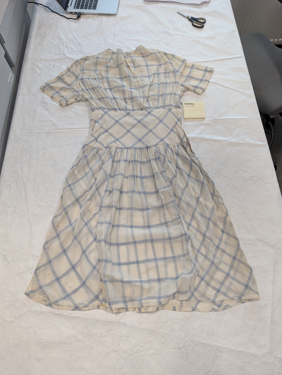 Hvit kjole med blå striper som lager et rutemønster. På overkroppen ligger rutene vertikalt, på midjen går stripene og rutene diagonalt og på skjørtet vertikalt og diagonalt i ulike felter. Kjolen har en hempe bak på kragen og en glidelåsa på siden under høyre arm.