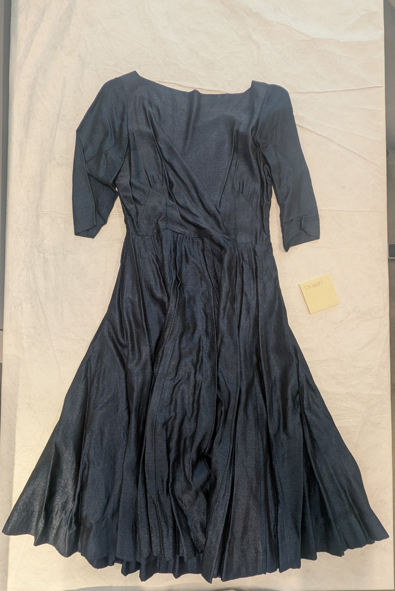 Blå kjole med halvlange armer, folder i skjørtet, skulderputer og glidelås på venstra side fra armhulen og ned. Det er vevd inn en blank tråd i stoffet som gjør at kjolen "skinner".