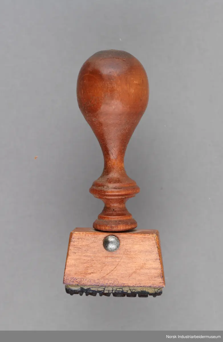 Stempelet har håndtak i lakkert tre med dreid profil. Håndtaket er naglet på stempelhodet.