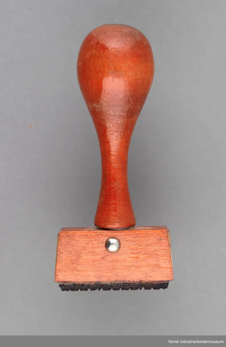 Stempelet har håndtak i lakkert tre med dreid profil. Håndtaket er naglet på stempelhodet.