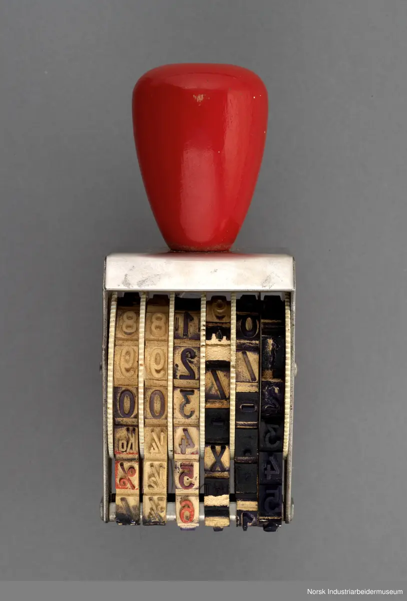 Stempelet i tre og metall. Det har seks bånd som kan roteres slik at det gir forskjellig trykk. Båndene har tall og noen bokstaver, tegn eller brøker.