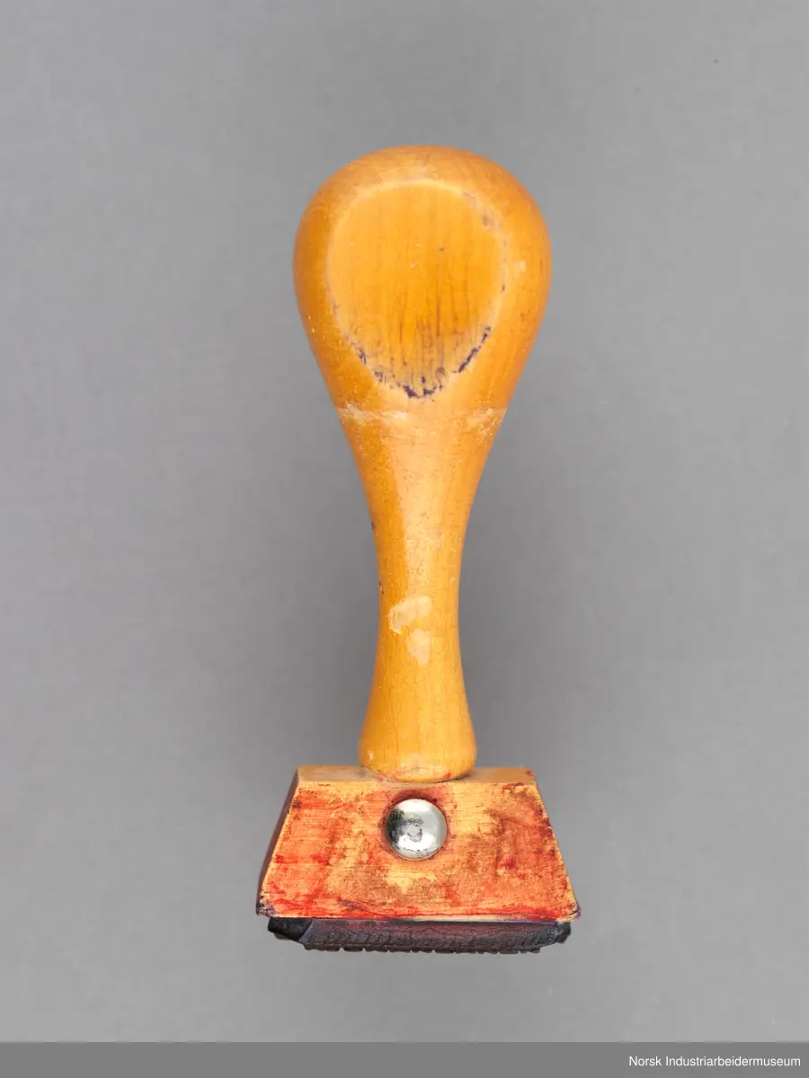 Stempelet har håndtak i lakkert tre som er naglet på stempelhodet. En siden av håndtaket er flatt.