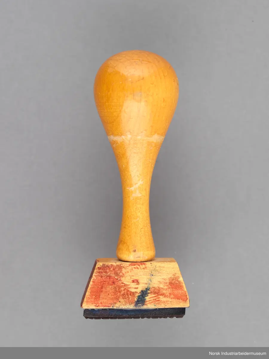 Stempelet har håndtak i lakkert tre som er naglet på stempelhodet. En siden av håndtaket er flatt.