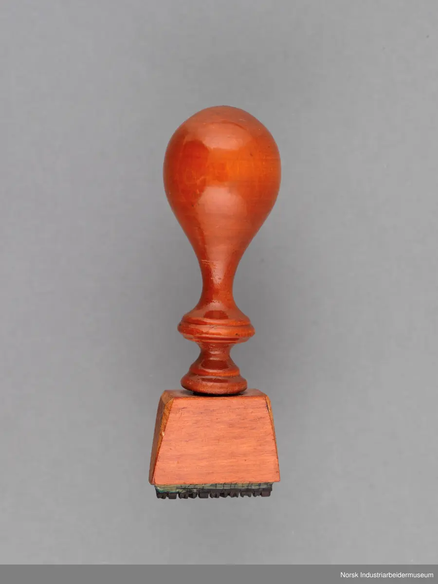 Stempelet har håndtak i lakkert tre med dreid profil. Håndtaket er naglet på stempelhodet.