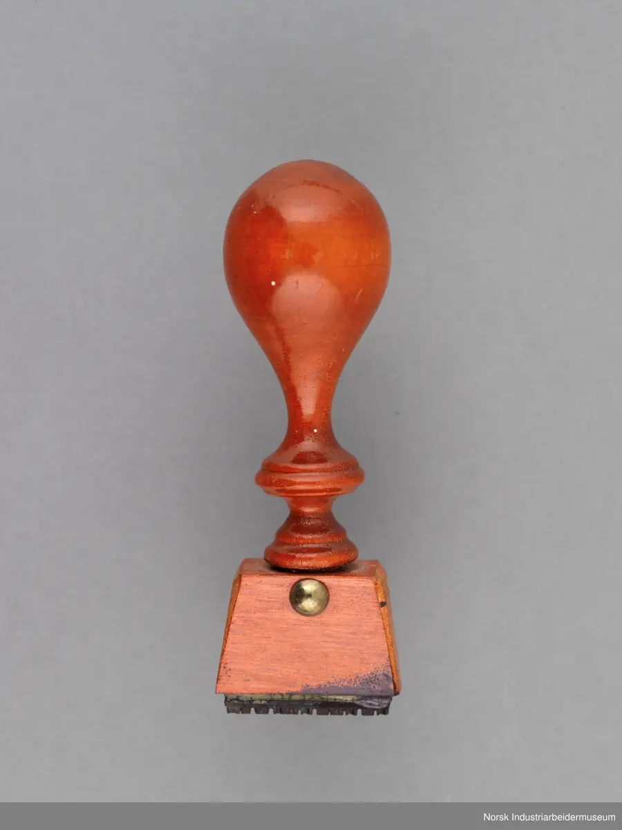 Stempelet har håndtak i lakkert tre med dreid profil. Håndtaket er naglet på stempelhodet.