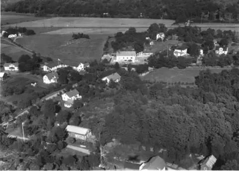 Flygbild över Kärradal. Bybebyggelse. Enfamiljshus och trädgårdar. Träd och buskar. Odlingslandskap. Vägar. Affärsbyggnad.