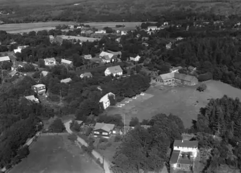 Flygbild över Kärradal. Bybebyggelse med enfamiljshus och trädgårdar. Lantgårdar med uthus och ekonomibyggnader. Träd och buskar. Odlingslandskap.