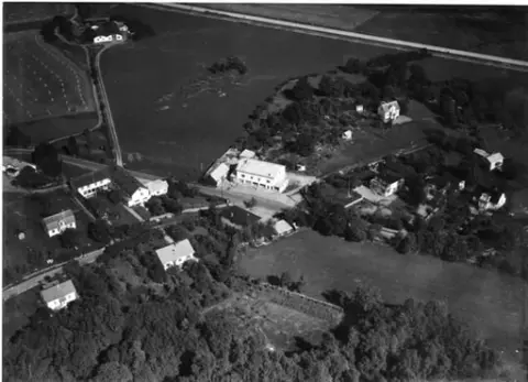 "Flygfoto över Kärradal. Sandows affär och Turisthotellet." Bybebyggelse med enfamiljshus och trädgårdar. Offentliga byggnader. Träd och buskar. Odlingslandskap.