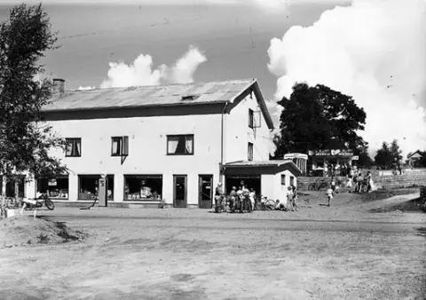 Affären i Kärradal, där en liten folksamling står och tittar på något i den lilla utbyggnadens i skyltfönster. Till höger ligger kiosk uppe på en höjd, där kunder köar.