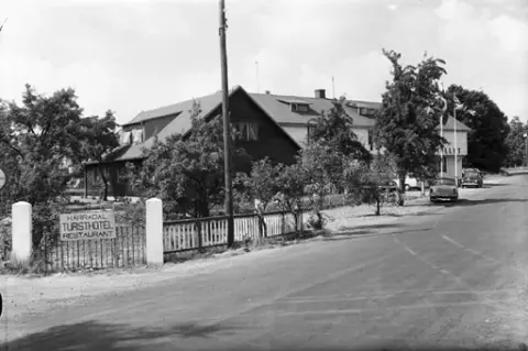 Turisthotellet i Kärradal . Framför hotellet står parkerade bilar. Hotellet omges av en trädgård och på grinden sitter en skylt med texten: "KÄRRADAL TURISTHOTEL RESTAURANT".