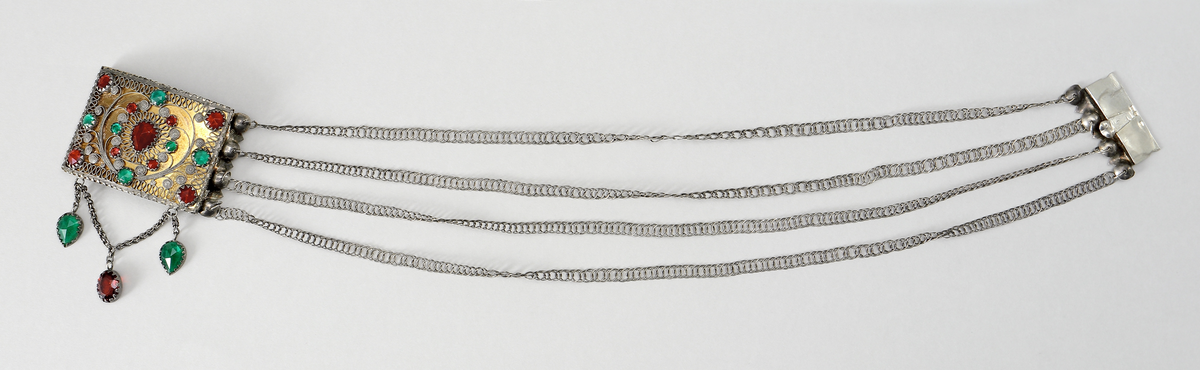 Halslås av silver, buren till värendsdräkt. Tillverkat som en välvd, grund dosa. på framsidan har den filigrandekor samt infattade röda och gröna fasetterade glasflusser. Bakgrunden är förgylld. På kortsidorna har den fyra tunna länkar, av pansartyp, fästade. Låsdelen består av en vikt, fjädrande silverplåt som skjuts in från sida. Under dosan hänger tre hänge bestående av två gröna och en röd slipad glasfluss. Dosans baksida är försedd med med ett broschspänne. Stämplar på låsets instickstunga.
Inskrivet i huvudkatalog 1968.