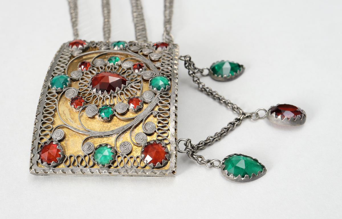 Halslås av silver, buren till värendsdräkt. Tillverkat som en välvd, grund dosa. på framsidan har den filigrandekor samt infattade röda och gröna fasetterade glasflusser. Bakgrunden är förgylld. På kortsidorna har den fyra tunna länkar, av pansartyp, fästade. Låsdelen består av en vikt, fjädrande silverplåt som skjuts in från sida. Under dosan hänger tre hänge bestående av två gröna och en röd slipad glasfluss. Dosans baksida är försedd med med ett broschspänne. Stämplar på låsets instickstunga.
Inskrivet i huvudkatalog 1968.