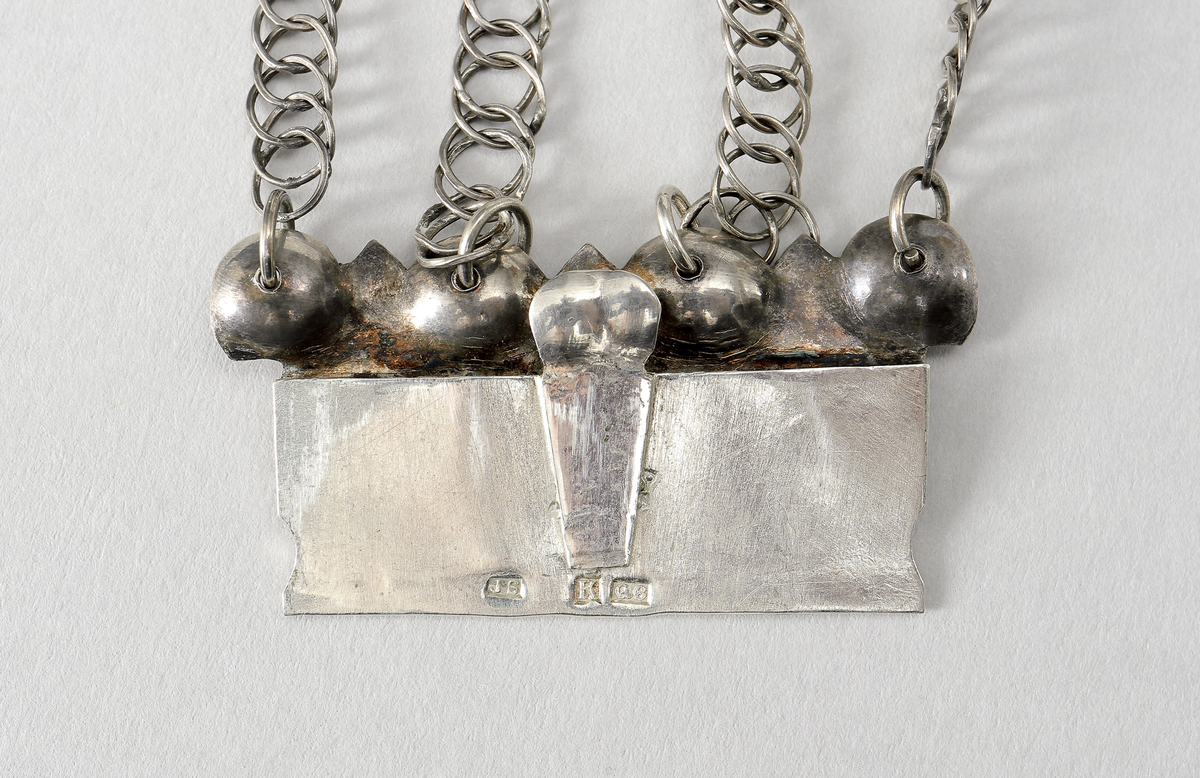 Halslås av silver, buren till värendsdräkt. Tillverkat som en välvd, grund dosa. på framsidan har den filigrandekor samt infattade röda och gröna fasetterade glasflusser. Bakgrunden är förgylld. På kortsidorna har den fyra tunna länkar, av pansartyp, fästade. Låsdelen består av en vikt, fjädrande silverplåt som skjuts in från sida. Under dosan hänger tre hänge bestående av två gröna och en röd slipad glasfluss. Dosans baksida är försedd med med ett broschspänne. Stämplar på låsets instickstunga.
Inskrivet i huvudkatalog 1968.