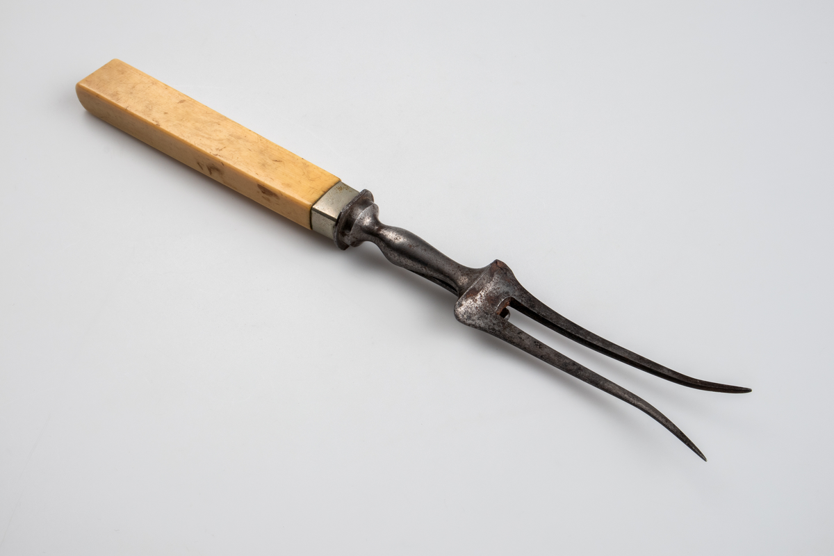 Gaffel i smidesjärn, med handtag av elfenben. Gaffeln har två klor som i den yttersta spetsen är utböjda och har ett smalt skaft av järn mellan klor och handtaget, på vilken ett utfällbart stöd är fästat. Handtaget är rektangulärt med avrundad ände. Runt handtagets nedre del är ett ca 1 cm brett band av silverplåt fästat. 
Funktion: Vid uppskärande av stekar.

Inskrivet i huvudboken 1903.