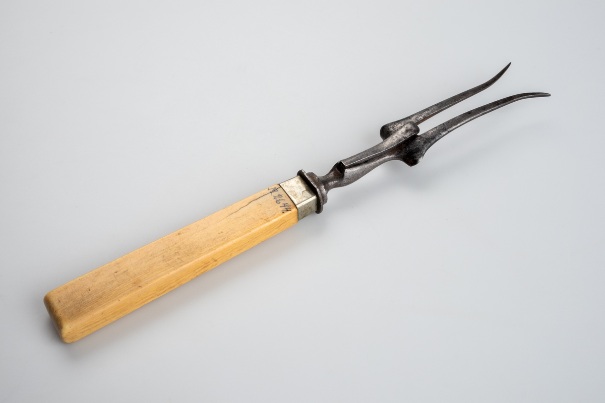 Gaffel i smidesjärn, med handtag av elfenben. Gaffeln har två klor som i den yttersta spetsen är utböjda och har ett smalt skaft av järn mellan klor och handtaget, på vilken ett utfällbart stöd är fästat. Handtaget är rektangulärt med avrundad ände. Runt handtagets nedre del är ett ca 1 cm brett band av silverplåt fästat. 
Funktion: Vid uppskärande av stekar.

Inskrivet i huvudboken 1903.