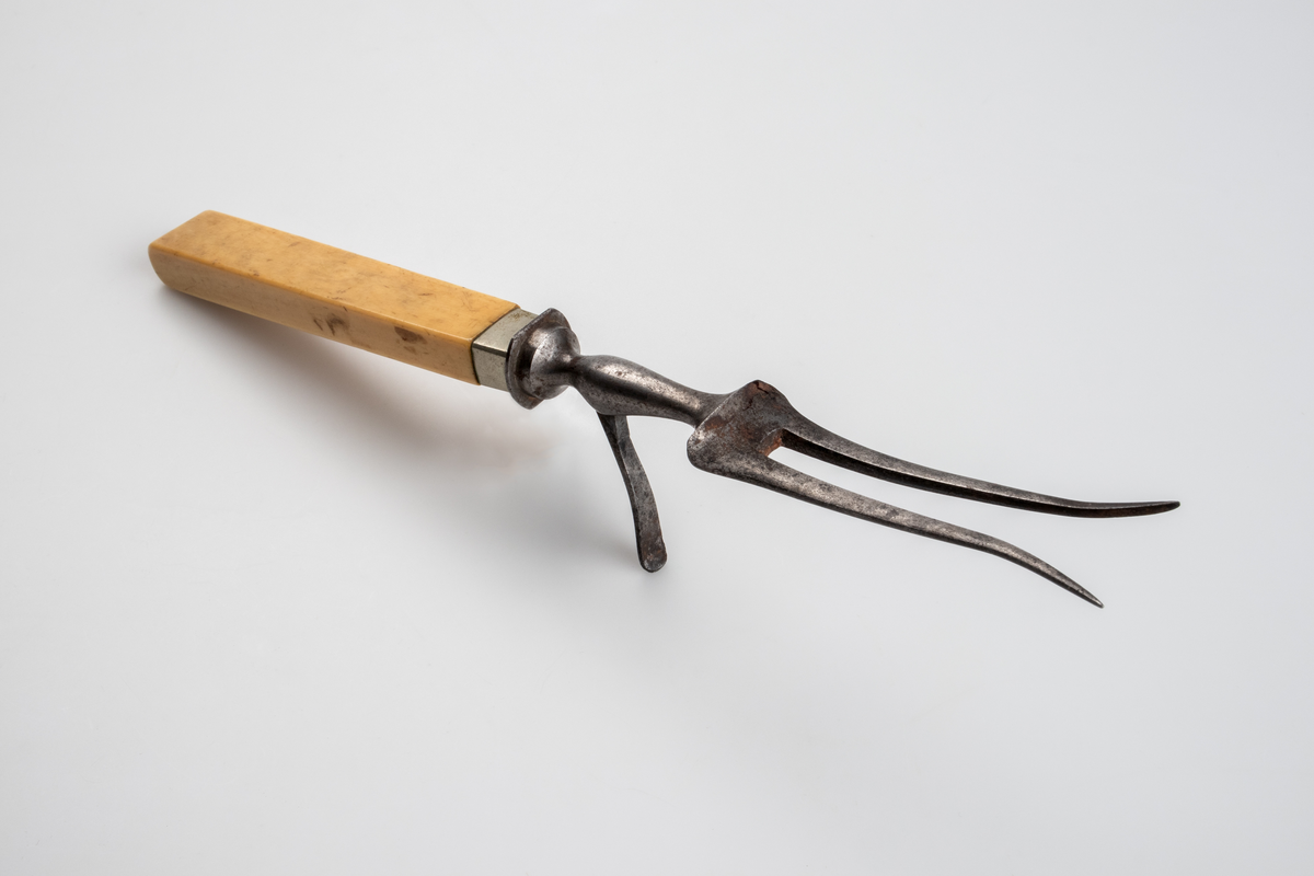 Gaffel i smidesjärn, med handtag av elfenben. Gaffeln har två klor som i den yttersta spetsen är utböjda och har ett smalt skaft av järn mellan klor och handtaget, på vilken ett utfällbart stöd är fästat. Handtaget är rektangulärt med avrundad ände. Runt handtagets nedre del är ett ca 1 cm brett band av silverplåt fästat. 
Funktion: Vid uppskärande av stekar.

Inskrivet i huvudboken 1903.