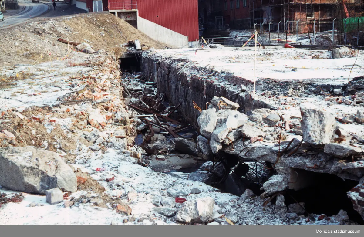 Bild tagen under 1970-talet. Fotografen har tagit bilden med Gamla Torget och Forsebron i ryggen. Fläckvisa snöpartier ligger utspridda och ses i solljuset.

Vi ser delar av den branta backen som i folkmun kallas för "Mölndals Kråka", samt trafik och en fotgängare i bakgrunden, i övre vänstra hörnet. 

I bakgrunden till höger ser vi grunden till SOAB:s kontorsbyggnad belägen i Stora Götafors och längre ned ser vi Nymans kvarn. I fokus så ser vi den stora rivningstomten och grundmuren till den fabriksbyggnad som tidigare har legat på platsen. Berol-Kemi har tidigare varit verksamma i den numera rivna byggnaden.
