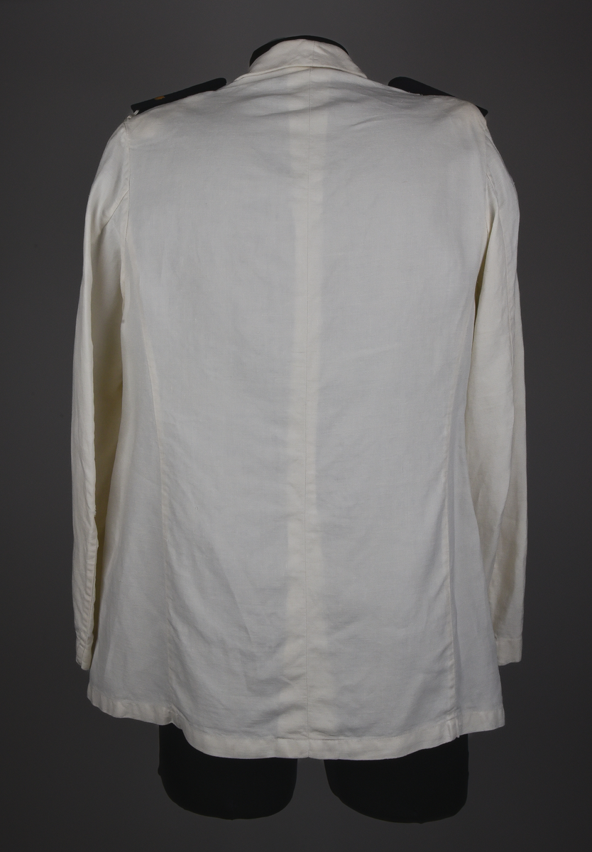 Kavaj av vitt linnetyg, dubbelknäppt med åtta knappar i brännförgylld gulmetall. Knappar enligt 1897-års reglemente. Framtill en snedställd bröstficka på vänster sida och två sidofickor. Löstagbara axelklaffar med gradbeteckning från år 1955 för bl.a. stationsmästare med tillhörande knappar från år 1953. Axelvaddar på insidan kavajen. Vid nacken hank för upphängning av kavajen. Rosa lapp från kemtvätt med text "GÄVLETVÄTTEN 1774 6/2" häftad vid hanken.
Kavajen tillhör 1921-års bestämmelser.