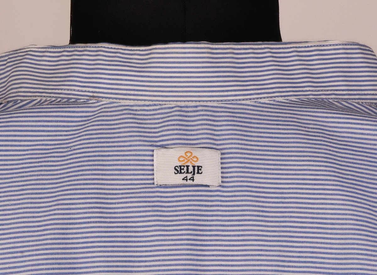 Långärmad skjorta i vit och ljusblå-randigt bomulls- och polyestertyg. Button down-skjorta med enkel knäppning med vita knappar. Bröstficka på vänster sida med maskinbroderad text "TX Logisitik" i rött garn ovanför fickan.
Etikett vid nacken text i svart: "SELJE 44".
Etikett vid vänster insida med text:
"Made by: SELJE
N-6740 SELJE 
67/33% Pol/Cot
Pr. No: 50301-1 
[symboler för tvättråd] Maskintvätt 60°, Klorblekmedel får ej användas, Varmt strykjärn, Endast kemtvätt med perkloreten, Får ej torktumlas".