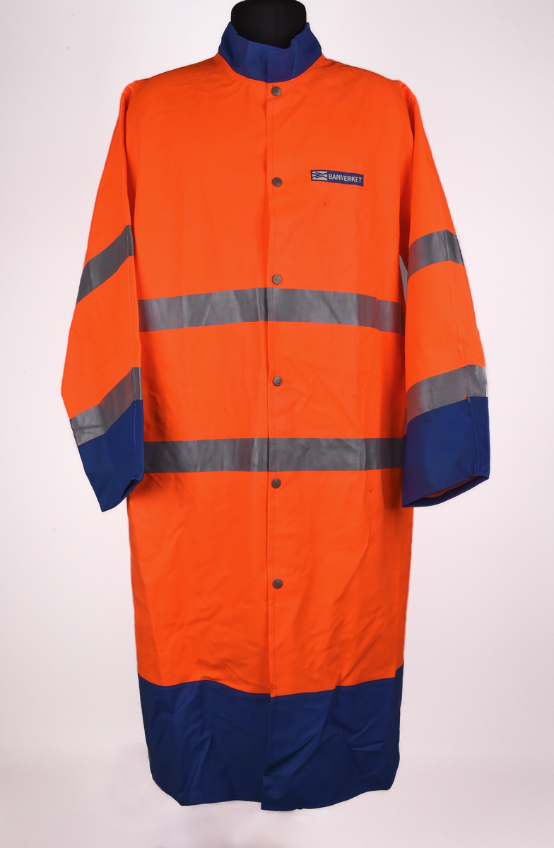 Orange regnrock med blå krage, ärmavslut och nedre kant. Rocken har två reflexband som går hela vägen runt, även runt ärmarna. Sex tryckknappar framtill. Banverkets logotyp i brösthöjd framtill på vänster sida och även på mitt på ryggen. 
Tre etiketter i nacken, med text: 
"This garment is made in GARDA-FLEX NX 64. The material is approved according to EN 533 Index 1. Detta plagg är tillverkat i GARDA-FLEX NX 64. Materialet är godkänt enligt EN 533 Index 1.", 
"XXL" och 
"CE 95 
EN 471 
prEN 343 
65% PVC 
35% POLYESTER 
B-G GARDA REGNKLÄDER AB
GÖTEBORGSV 175 502 60 BOR
www.gardagarments.se
[Symboler för tvättråd:] Maskintvätt 40°, Klorblekmedel får ej användas, Får ej strykas, Ej kemtvätt, Får ej torktumlas
max 25x
[Storleksguide vid illustration av människa:] byst 116-124, midja 84-88, kroppslängd 175-188".