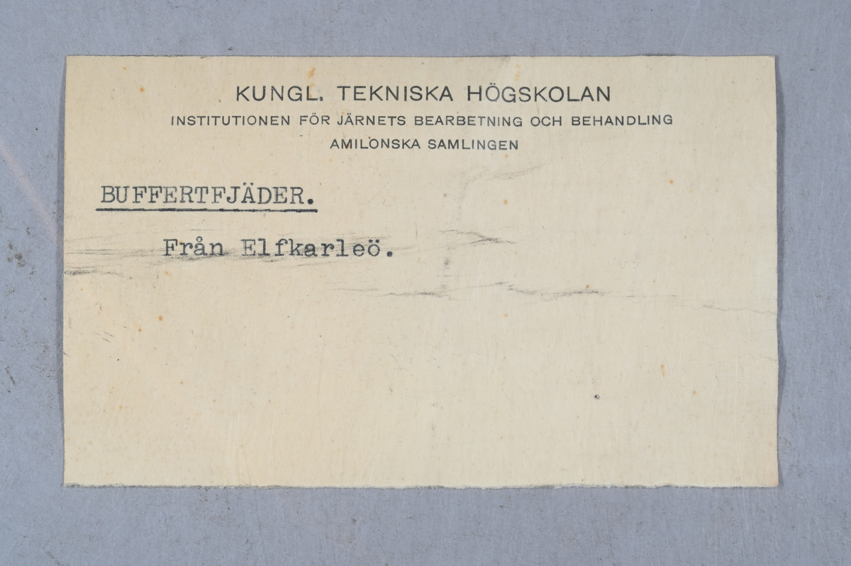 Prov på buffertfjäder, svart. Från Elfkarleö bruk. Tillhörande lapp med samma notering. Märkning på provet med påklistrad etikett med notering: "225". Märkning på provet med notering: "Elfkarleöbruk".