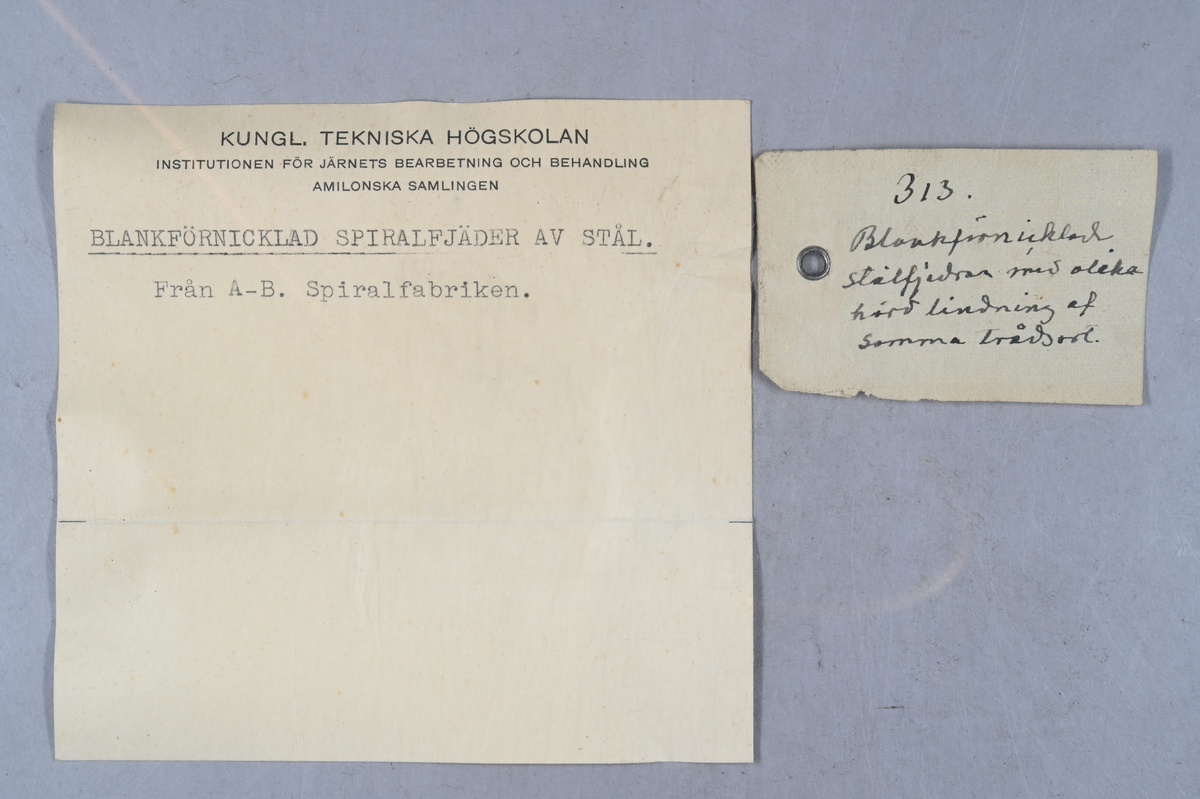 Två prover på blankförnicklad spiralfjäder av stål. Tillverkade av A-B. Spiralfabriken. Tillhörande lapp med samma notering. Tillhörande etikett med notering: "313. Blankförnicklad stålfjäder med lika hård lindning af samma trådsort."
