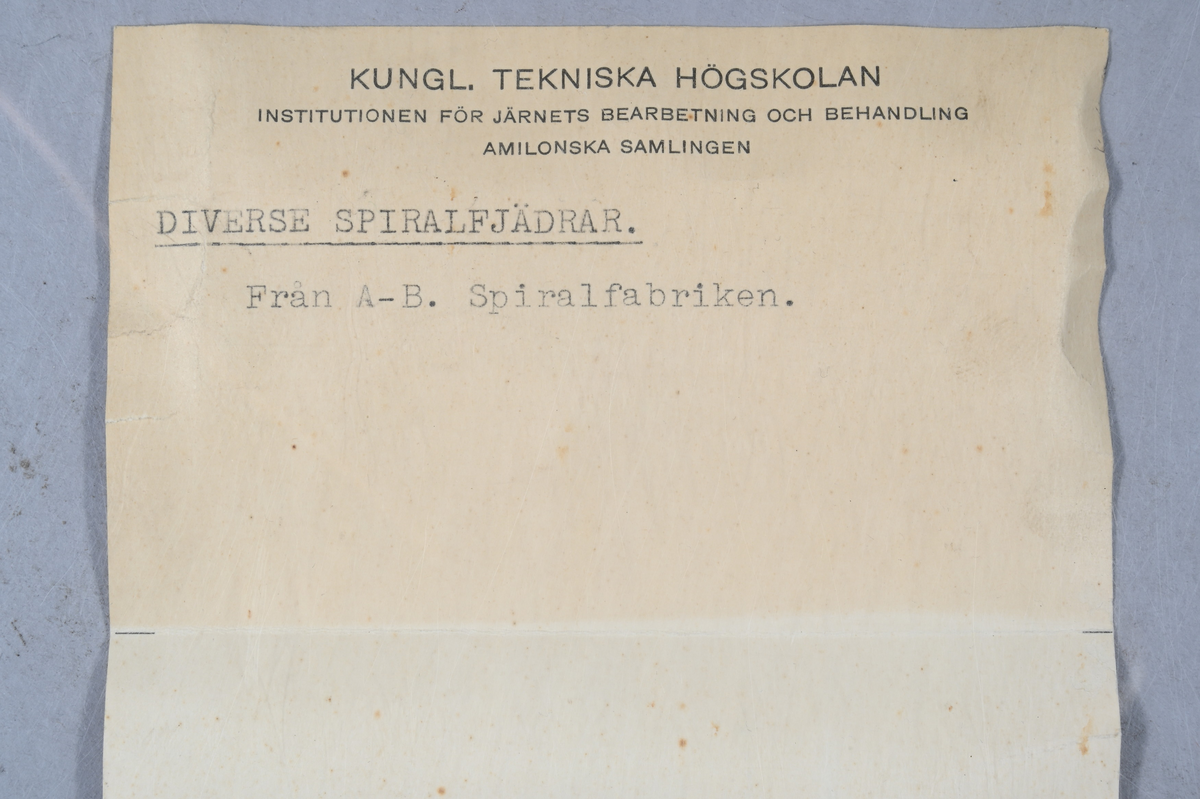 Två prov på diverse spiralfjädrar, svart. Tillverkad av A-B. Spiralfabriken. Tillhörande lapp med samma notering.
