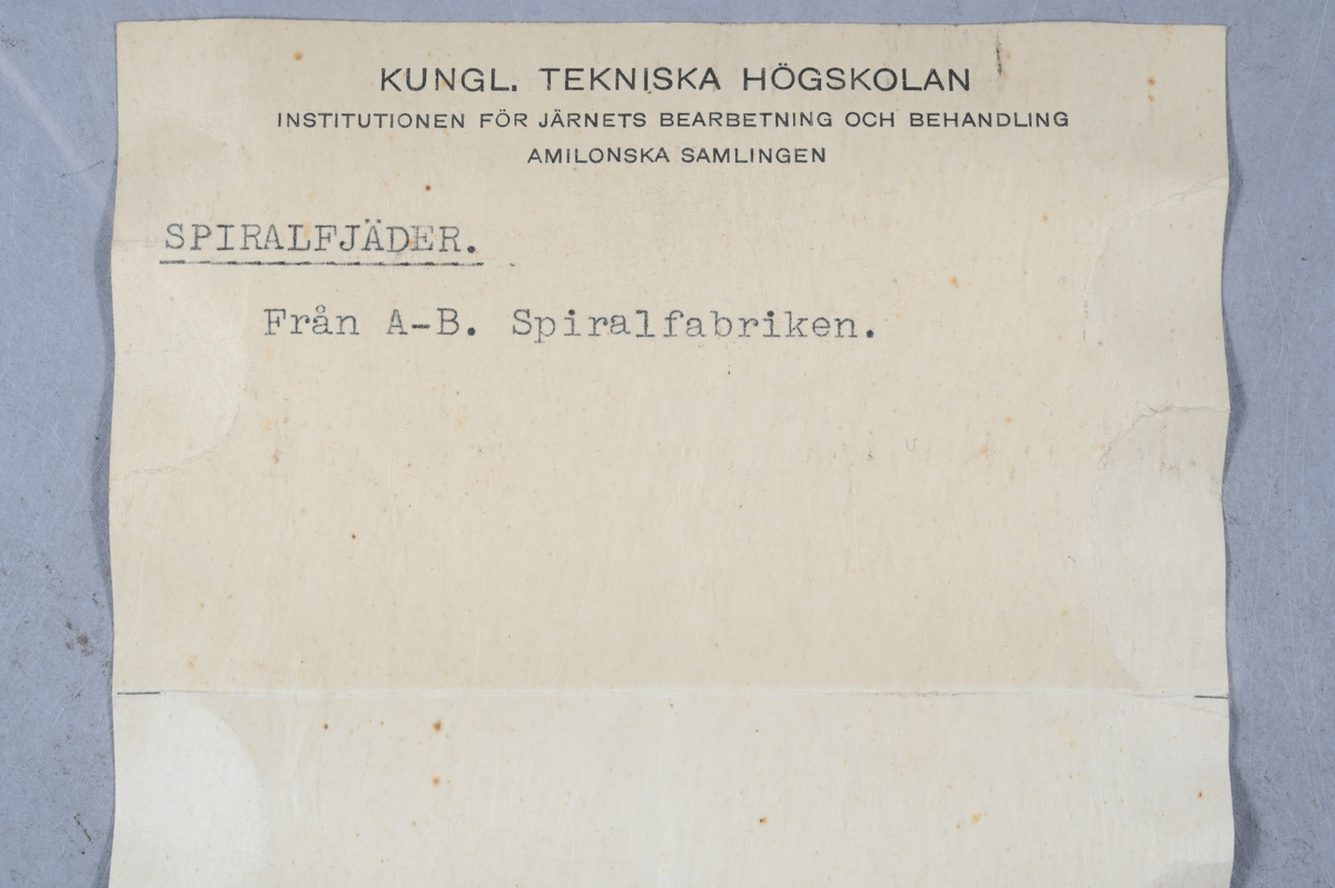 Prov på spiralfjäder, svart. Tillverkad av A-B. Spiralfabriken. Tillhörande lapp med samma notering.