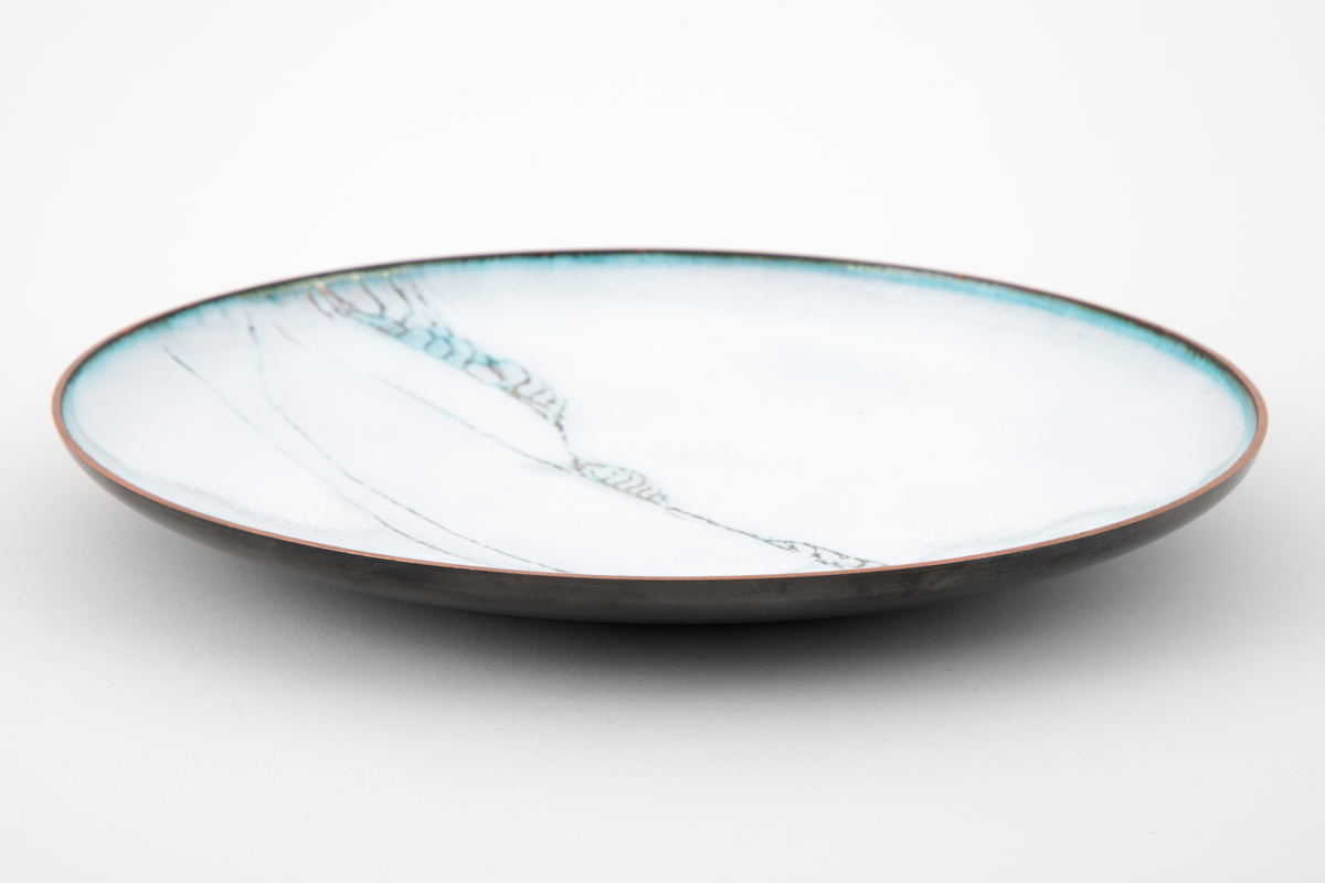 Rundt fat i kobber. Fatet er svakt konkavt. Oversiden er dekket av hvit emalje med et spinkelt linjemønster i gull, som enkelte steder går over i turkis. Turkis også langs munningskanten.