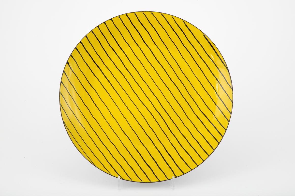 Sirkelrundt fat i matt emalje på stål. Oversiden gul med parallelle, uregelmessige sorte linjer. Undersiden sort.