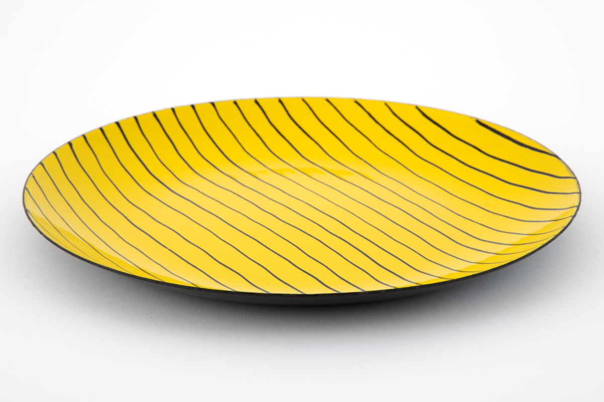 Sirkelrundt fat i matt emalje på stål. Oversiden gul med parallelle, uregelmessige sorte linjer. Undersiden sort.