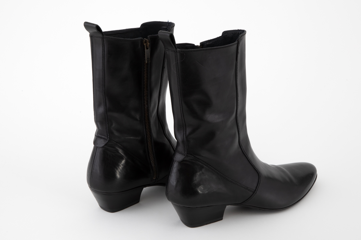 Relativt høye ankelstøvletter av svart lær. Spiss,rundet tå, Kraftig metallglidelås på innsiden. Tåkappe og overlær går iett og smalner til en stolpe midt foran, ca 1 cm høyere enn resten av støvelskaftet. Tilsvarende, meget smal stolpe, er påsydd midt bak. Den har en løkke øverst til å trekke i når støvelen skal på, og ender i overkant av hælkappen. Alle sømmer er stukne, med to tettstilte sømmer. Relativt høy, kraftig hæl, som skråner kraftig innover. Helfôret med midnattblått skinn. Tynn lærsåle uten mønstring. Flikken er sammensatt, med bakre del pålimt og fremre del stiftet med tettstilte stifter. Innvendig såle er naturfarget, men pålimt et svart stykke tynt skinn under hæl og gelenk. Str. 44.