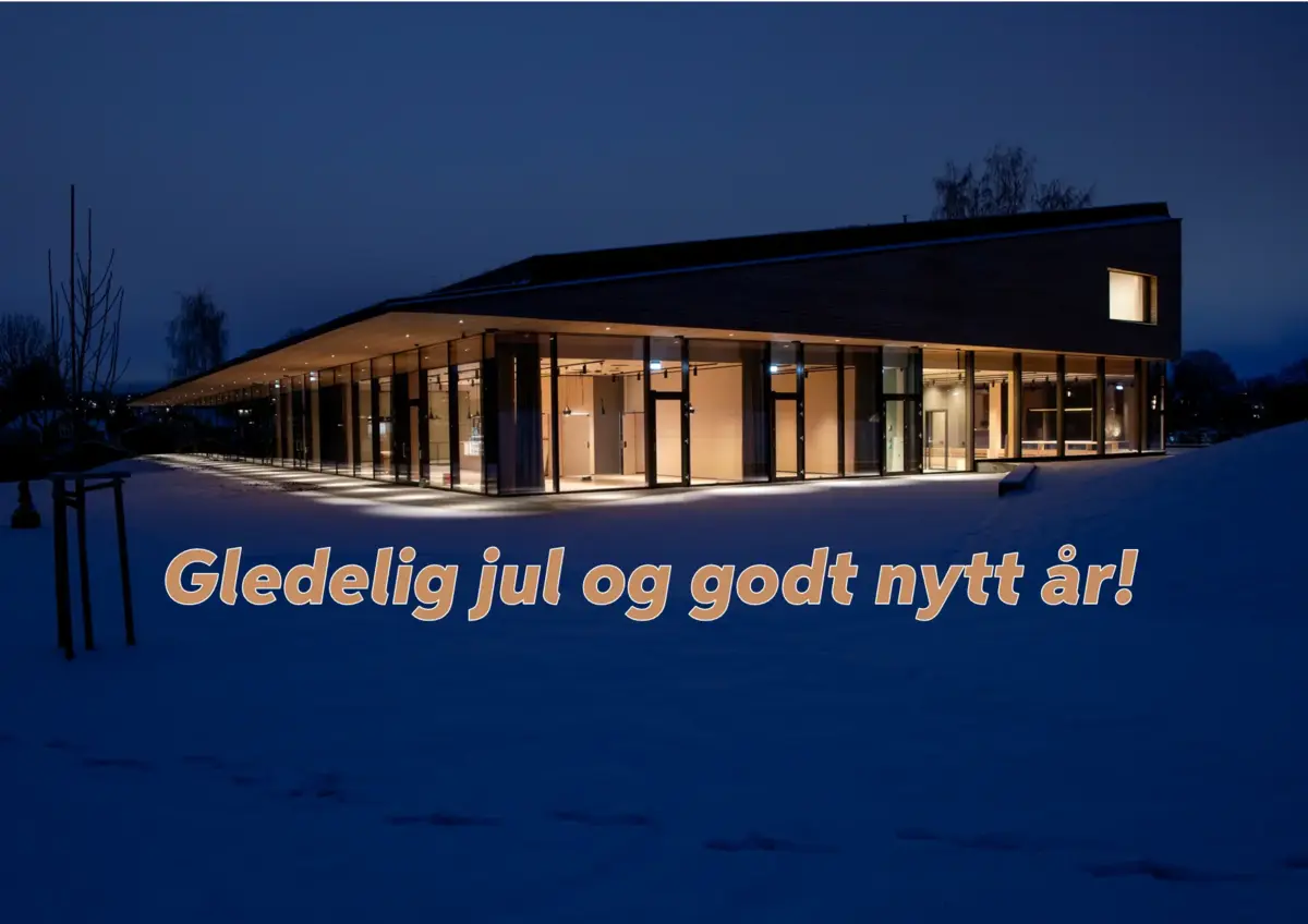 Bilde av Anno Domkirkeodden historiesenter som lyser opp i kveldsmørket, med snø på bakken rundt. Bildet har påskrevet teksten "Gledelig jul og godt nytt år!"