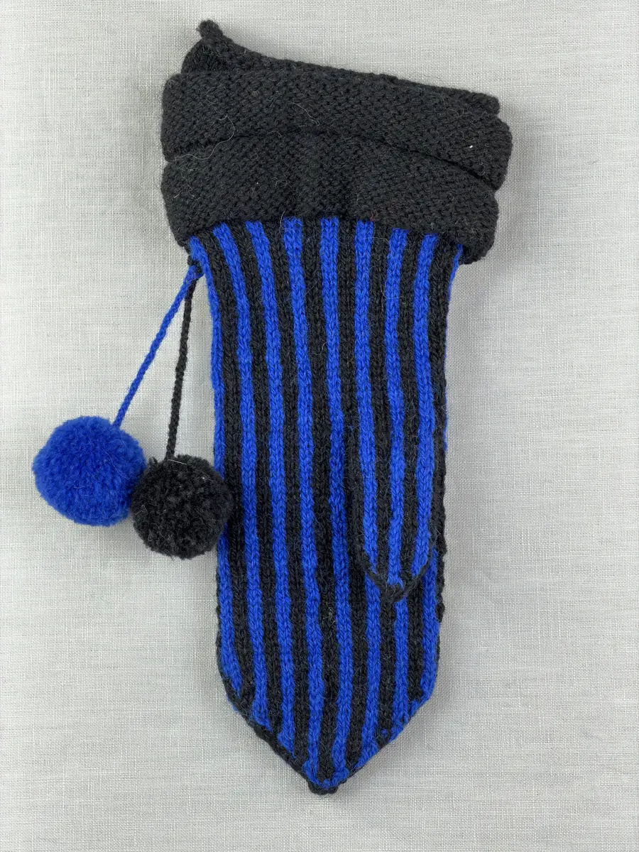 En stickad tumvante, med randad mudd/krage i slätstickning och vante i tvåfärgad slätstickning, av 2-trådigt ullgarn. Vanten har två garnbollar med påtad snodd och bandhoptagning i toppen.
