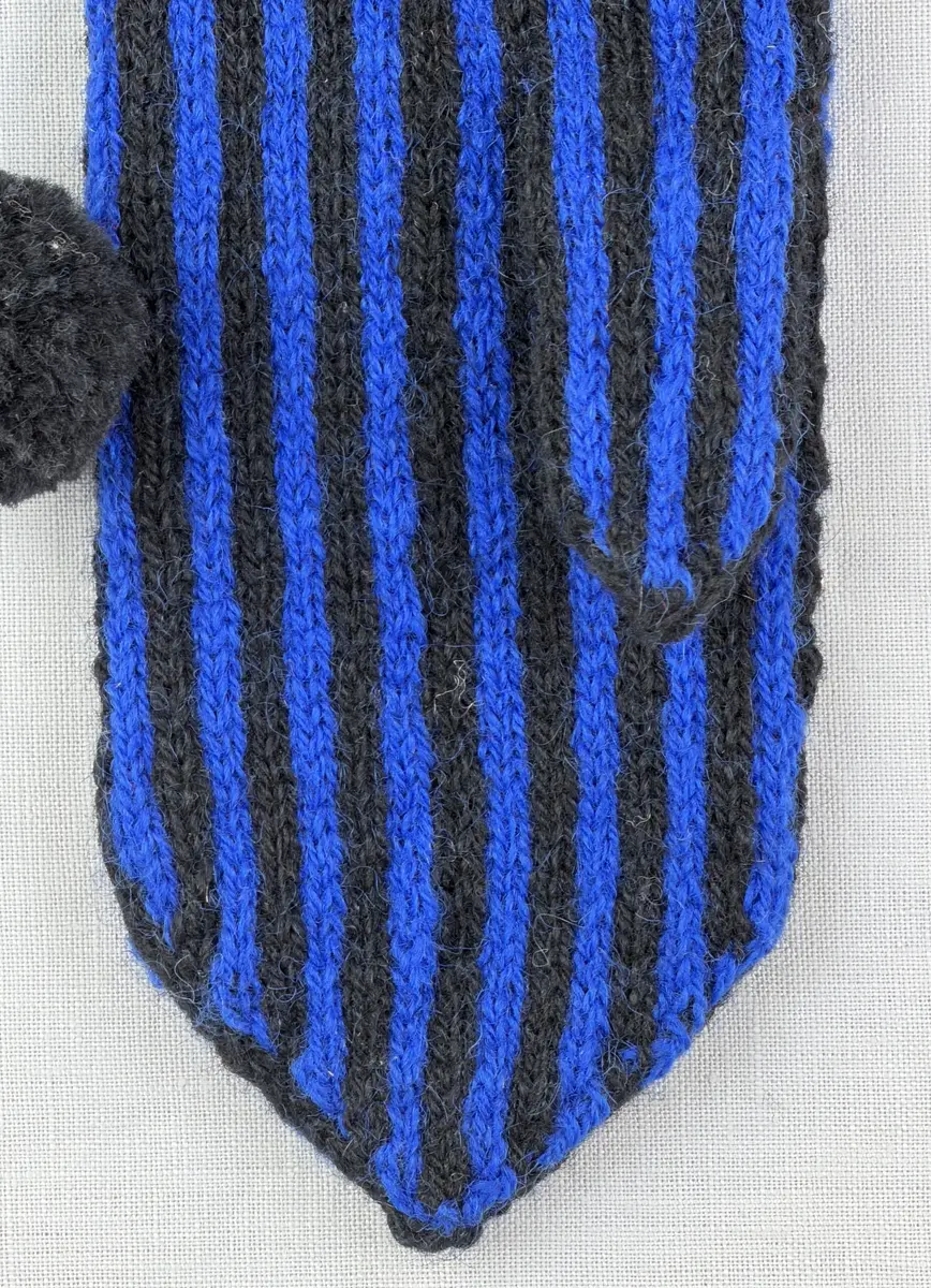 En stickad tumvante, med randad mudd/krage i slätstickning och vante i tvåfärgad slätstickning, av 2-trådigt ullgarn. Vanten har två garnbollar med påtad snodd och bandhoptagning i toppen.
