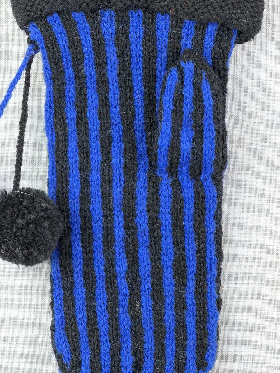 En stickad tumvante, med randad mudd/krage i slätstickning och vante i tvåfärgad slätstickning, av 2-trådigt ullgarn. Vanten har två garnbollar med påtad snodd och bandhoptagning i toppen.
