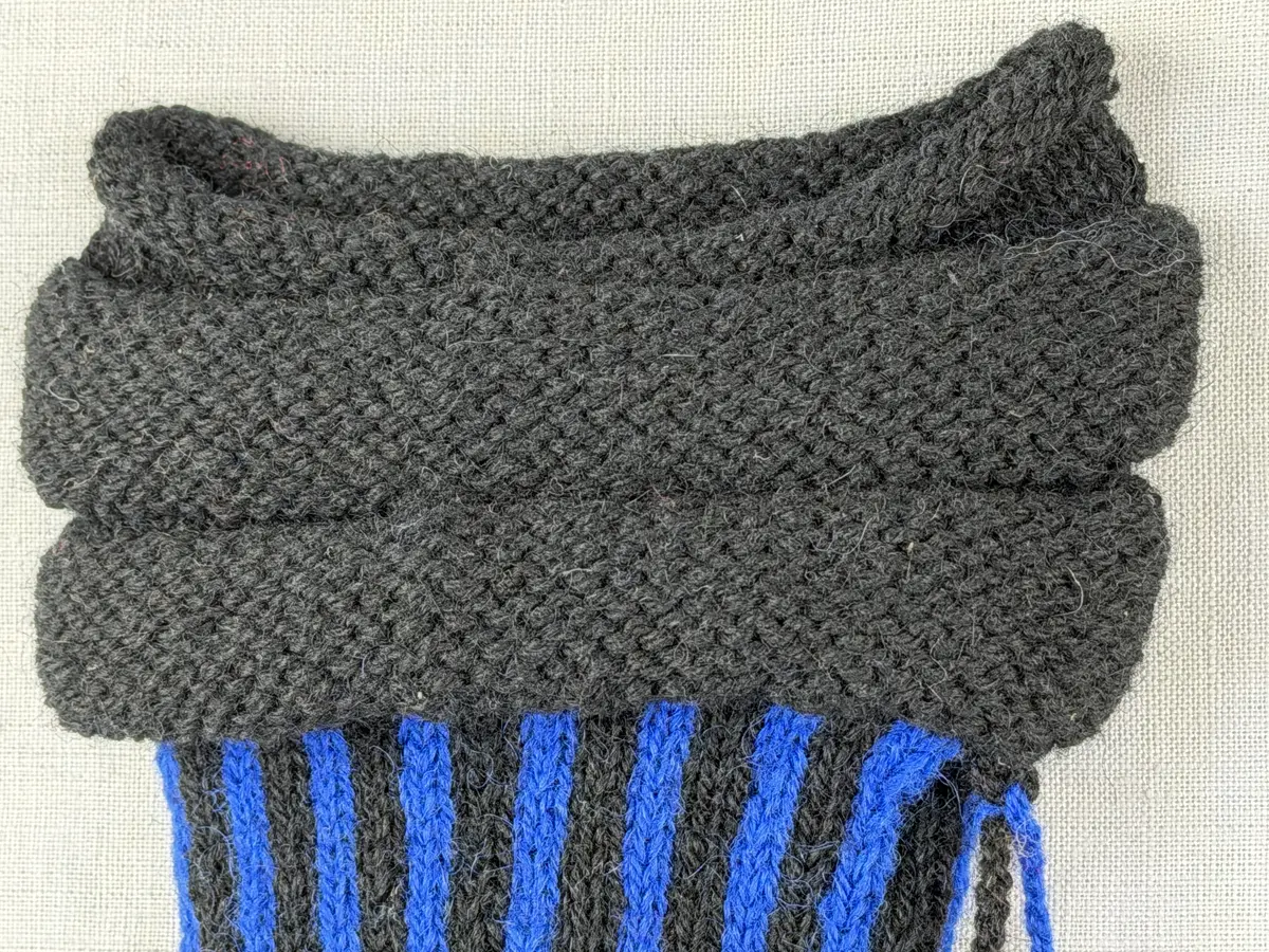 En stickad tumvante, med randad mudd/krage i slätstickning och vante i tvåfärgad slätstickning, av 2-trådigt ullgarn. Vanten har två garnbollar med påtad snodd och bandhoptagning i toppen.
